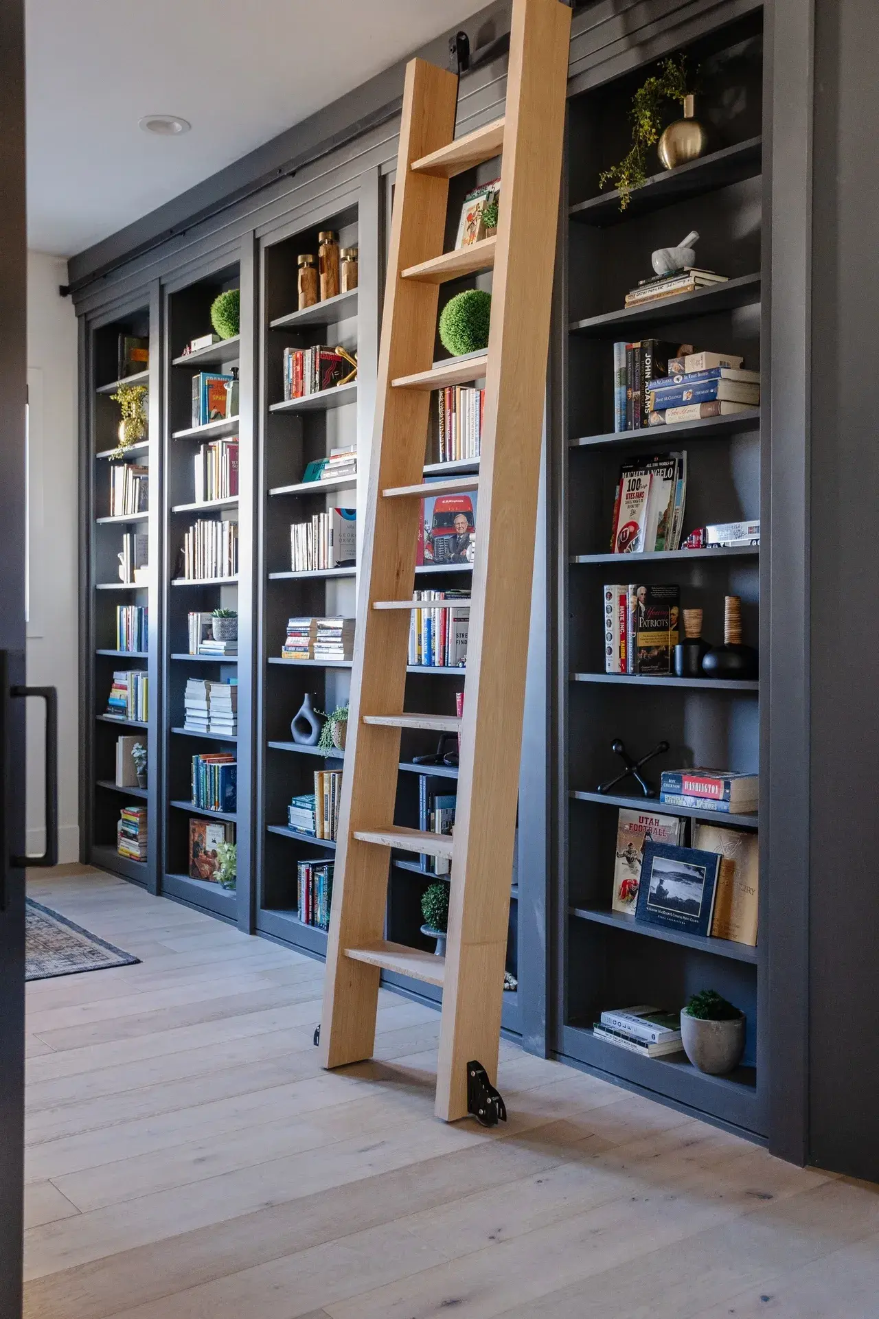 Sophisticated home library with custom cabinetry, dark shelving, and a rolling wooden ladder in a luxury custom home in Draper, Utah by E Builders