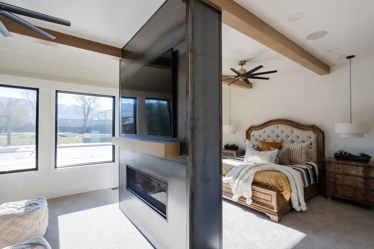 Spacious primary bedroom with a cozy double-sided fireplace and custom TV wall in a luxury custom home in Draper, Utah by E Builders